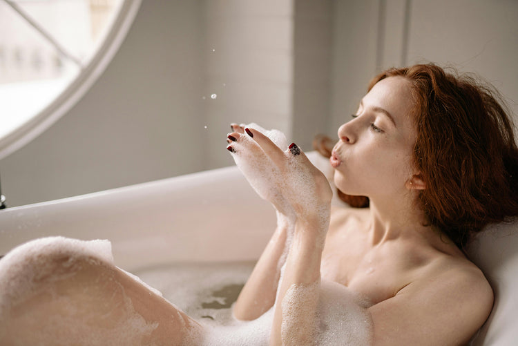 Woman relaxing in a bubble bath with eyes closed, wearing moisturizer