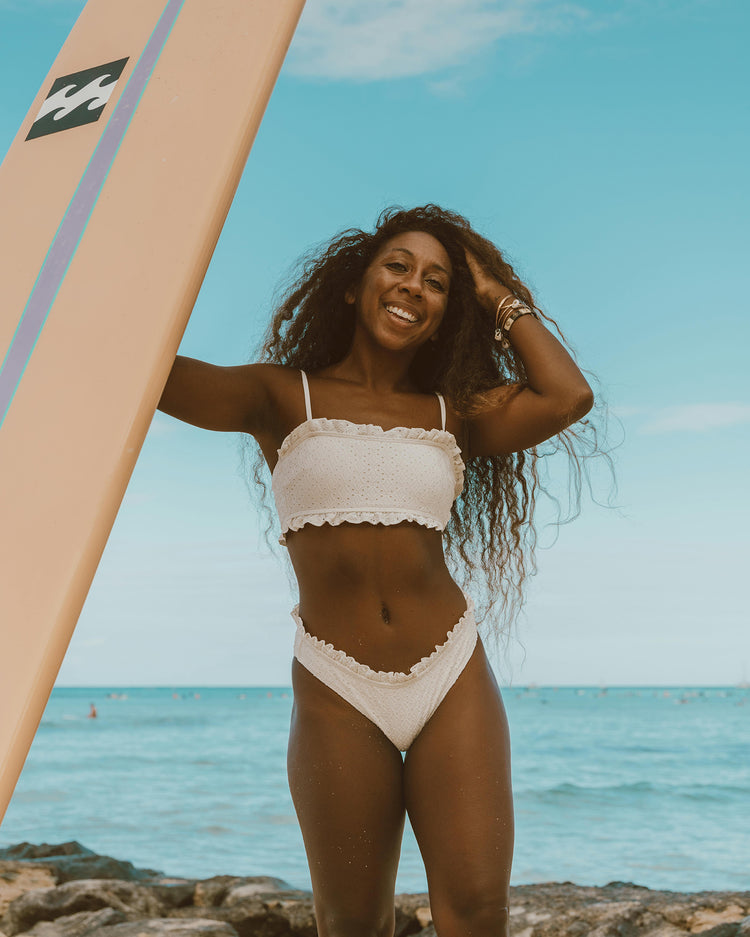 Woman in a bikini standing on a beach with a surfboard, smiling, moisturizer.