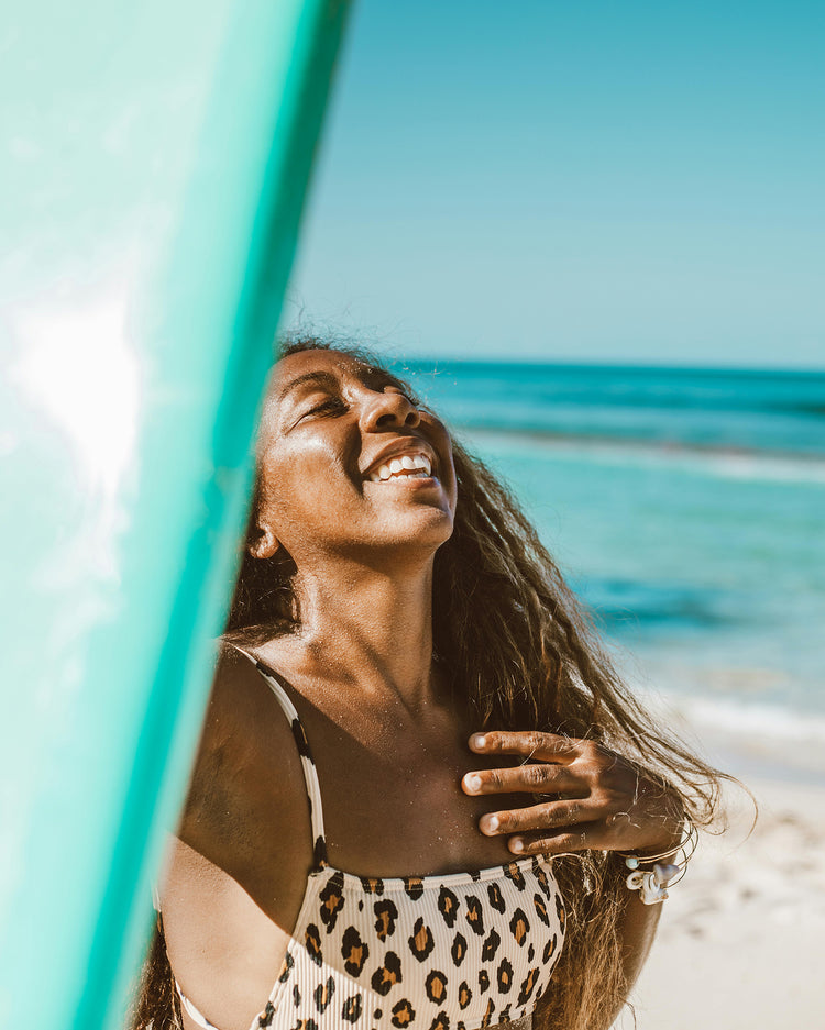 Woman wearing moisturizer in a leopard print dress standing on a beach with turquoise water and sky.